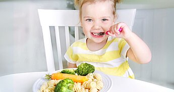 Malé dítě sedí u stolu, v ruce drží lžíci a před sebou má talíř se zeleninou a těstovinami.