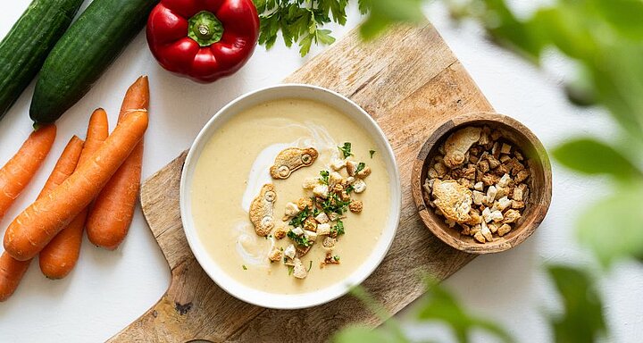 Misku krémové polévky zdobené krutony a bylinkami doplňuje čerstvá zelenina na dřevěném prkénku.