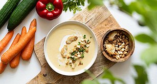 Misku krémové polévky zdobené krutony a bylinkami doplňuje čerstvá zelenina na dřevěném prkénku.