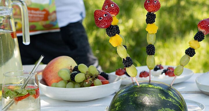Na stole je připraven meloun, ovocné špízy, sklenice vody s brčkem a mísa s hrozny a dalším ovocem.