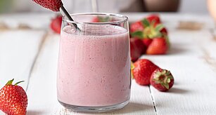 Sklenice narůžovělého koktejlu s jahodou na kovovém brčku, obklopená jahodami na bílém dřevěném povrchu.