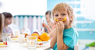 Dieťa v modrej košeli jedáva plátok pomaranča, pred sebou má tanier s ovocím a nápojmi, zatiaľ čo ostatné deti sedia pri stole v pozadí.