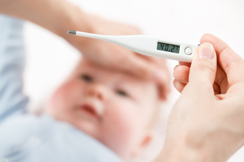 Ruka drží digitální teploměr, který ukazuje teplotu 40,2 °C, na rozmazaném pozadí je obličej dítěte.