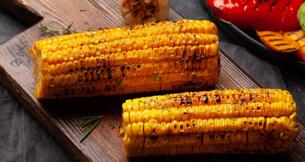 Grilovaná kukuřice na klasu se spálenými skvrnami a ozdobená bylinkami, servírovaná na dřevěném prkénku.