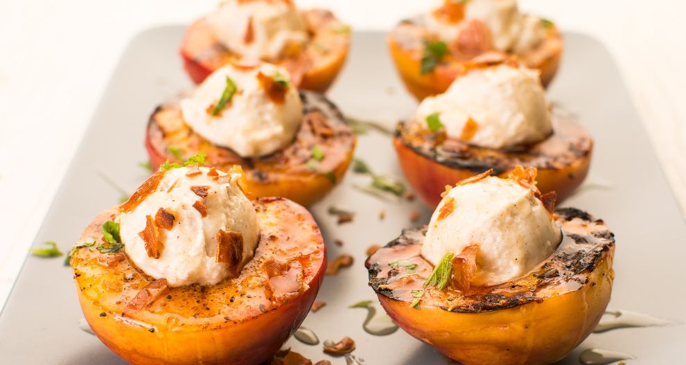 Grilované půlky broskví přelité krémovou hmotou, ozdobené bylinkami a kousky slaniny.