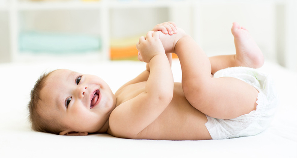 A baby lies on a white surface, holding its foot.