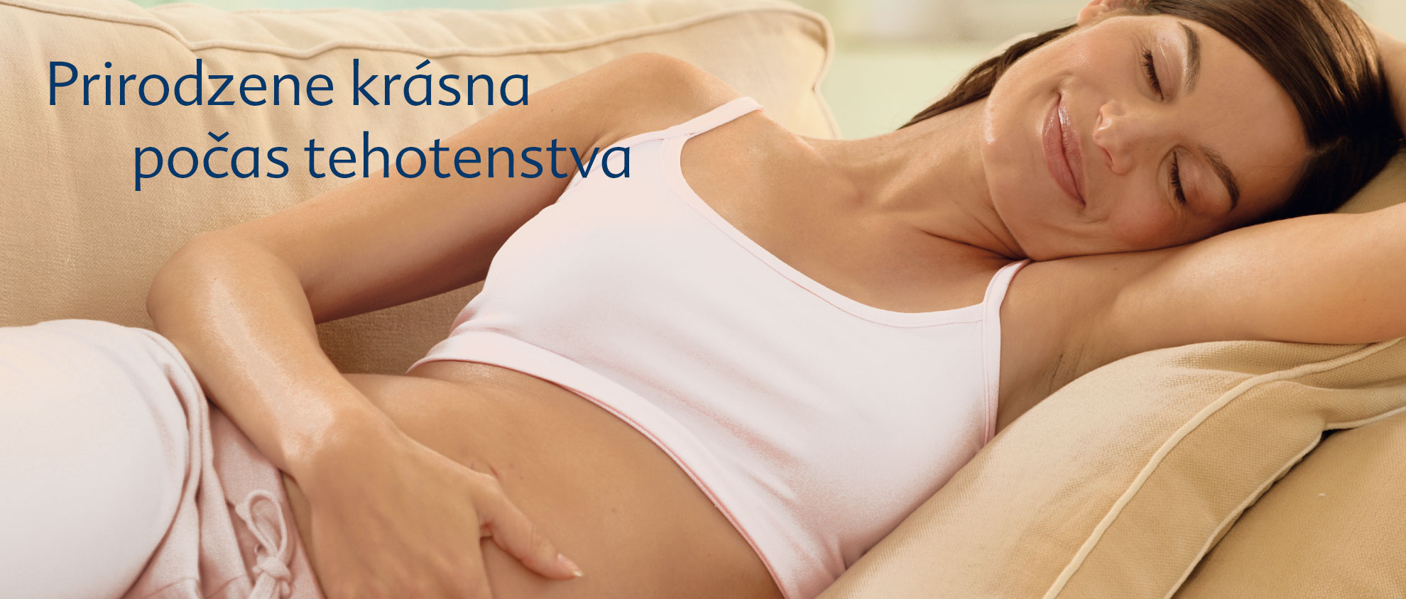 Žena v bielom tielku a svetlých nohaviciach leží na béžovom gauči, ľavou rukou si podopiera hlavu a pravú ruku má položenú na bruchu.