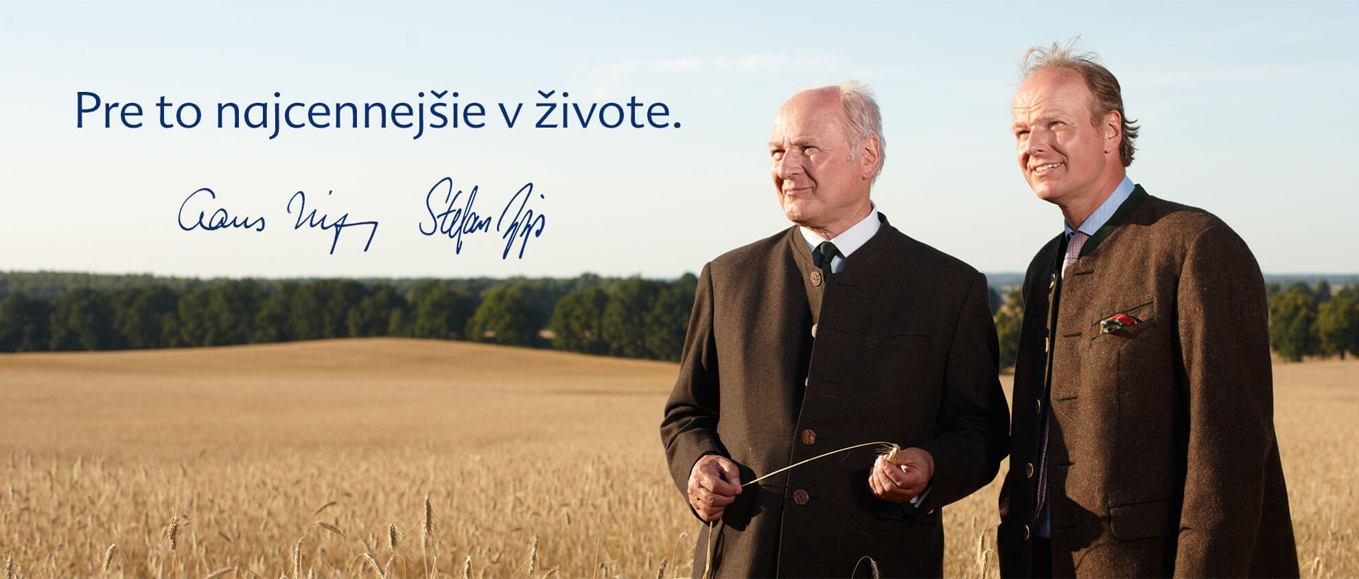 Dvaja muži v oblekoch stoja na pšeničnom poli s malebným pozadím stromov a jasnej oblohy.