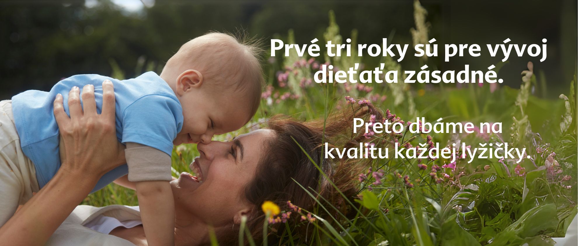 Žena a dieťa ležia na tráve obklopené kvetmi a žena sa pozerá na dieťa.