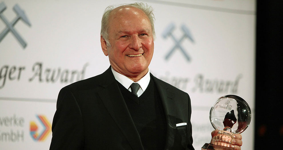 Starší muž v čiernom obleku stojí na pódiu a v ruke drží priehľadné guľovité ocenenie s nápisom "Schweizer Award" a logami v pozadí.