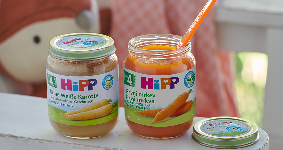 Dve sklenené nádoby s oranžovým a hnedým obsahom, označené nápisom "HIPP", sú umiestnené na bielom povrchu.