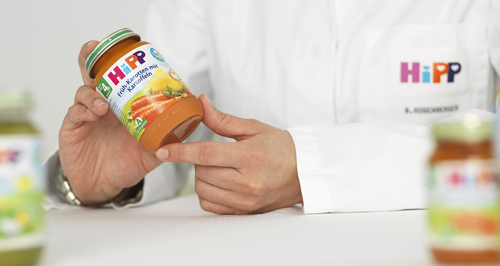 Osoba v bielom plášti drží nádobu s detskou výživou s nápisom "HiPP" a "Karotten-Sauce mit Apfel" (Mrkvová omáčka s jablkom).