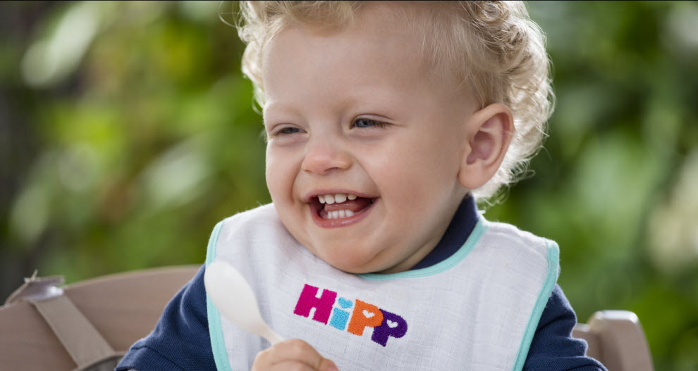 Malé dieťa s kučeravými vlasmi má na sebe podbradník s nápisom "HIpp" a v ruke drží lyžičku.