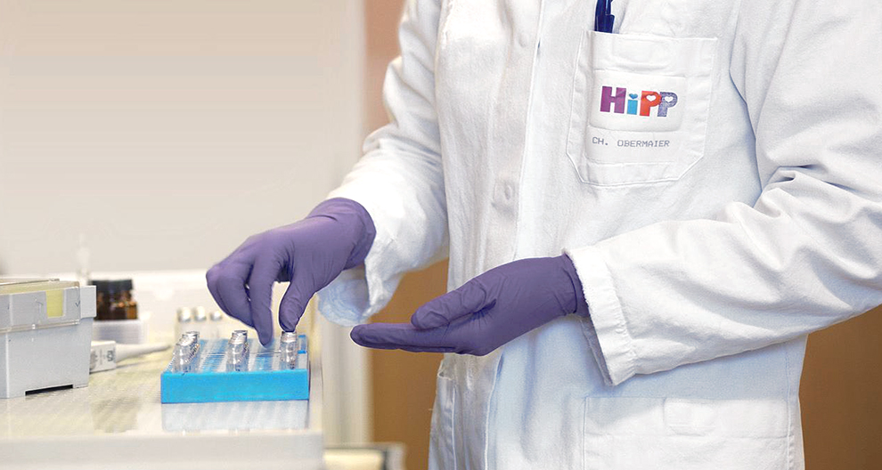 Osoba v bielom laboratórnom plášti s logom "HiPP" a názvom "CH. OBERMAIER" manipuluje s modrým podnosom s malými fľaštičkami.