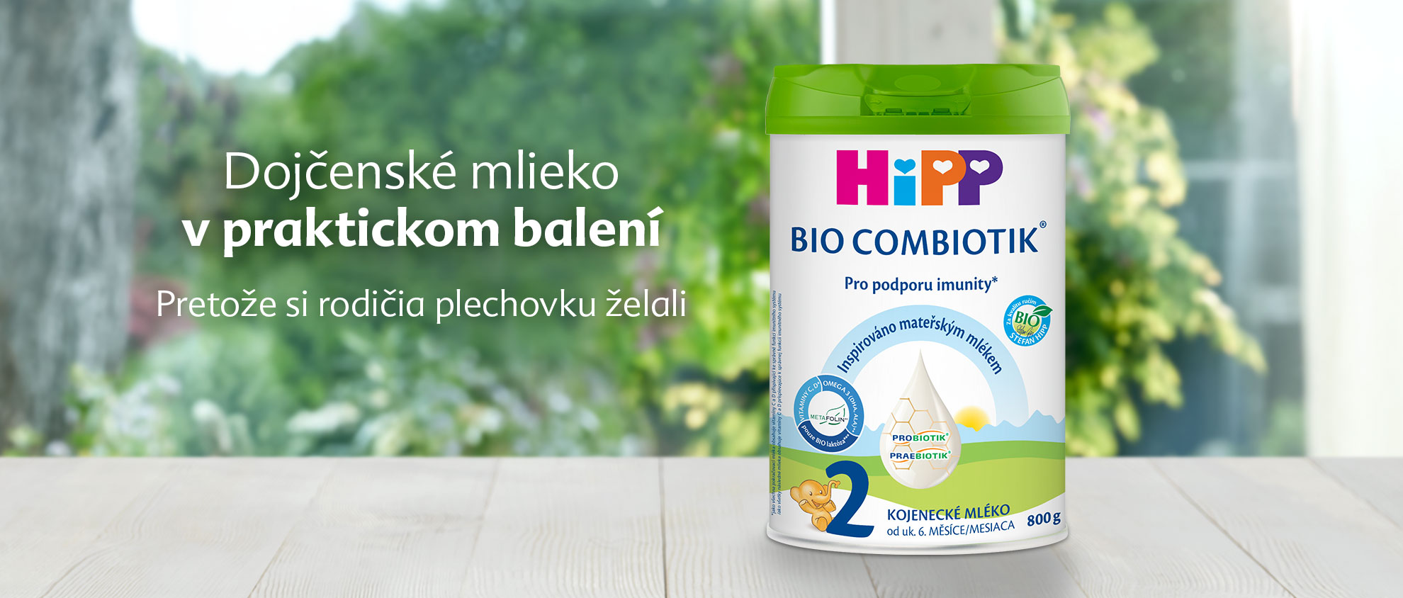 Biela valcovitá nádoba so zelenými a modrými štítkami s ilustráciou dieťaťa a textom "Hipp Bio Combiotik" je umiestnená na drevenom povrchu na rozmazanom pozadí zelene.