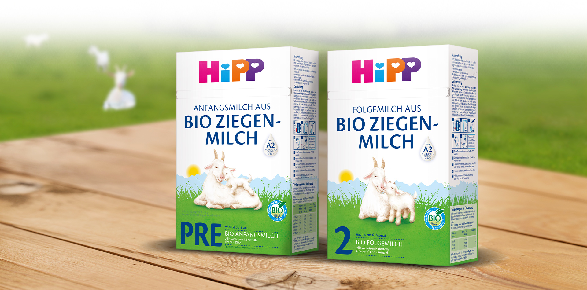 Dve biele škatule s farebným textom a ilustráciami kozy a kozliatka sú umiestnené na drevenom povrchu s rozmazaným zeleným pozadím s bielou kozou.