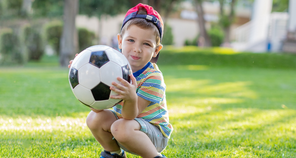 Chlapec vo farebnom pruhovanom tričku a červenej čiapke drží na trávnatom ihrisku futbalovú loptu.