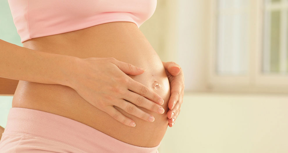 V interiéri sedí tehotná žena v ružovom oblečení s rukami jemne položenými na bruchu.