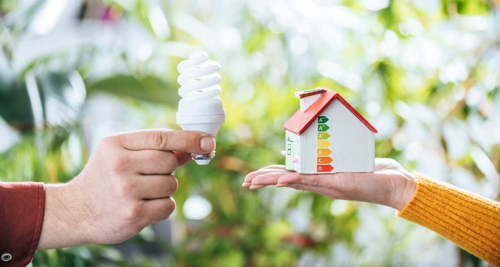 Ruka drží špirálovú kompaktnú žiarivku, zatiaľ čo druhá ruka predstavuje model domu s hodnotením energetickej účinnosti.