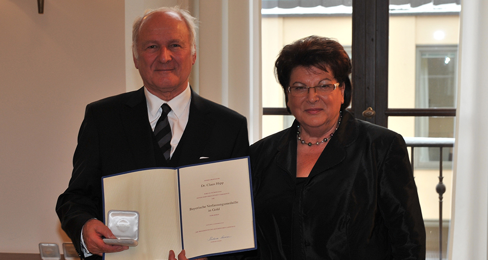 Starší muž v čiernom obleku drží certifikát a okrúhly strieborný predmet, stojí vedľa staršej ženy v čiernom oblečení, obaja pózujú v interiéri s oknom v pozadí.