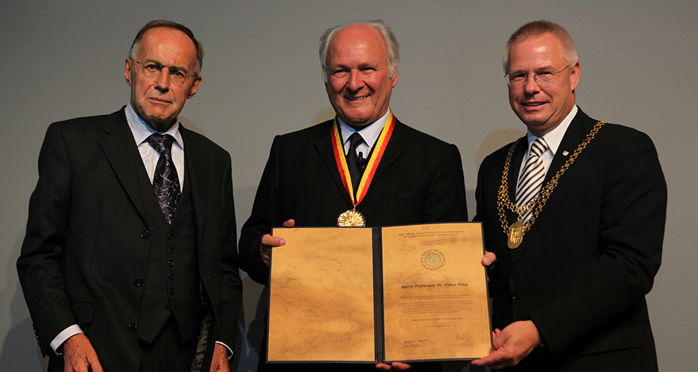 Traja muži vo formálnom oblečení stoja spolu, pričom ústredná postava drží veľký, zdobený certifikát a medailu.