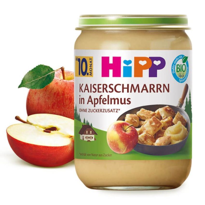 Pohár s detskou výživou s etiketou v nemčine, na ktorej je vyobrazené jablko a miska s jedlom, je položený vedľa celého a nakrájaného jablka.